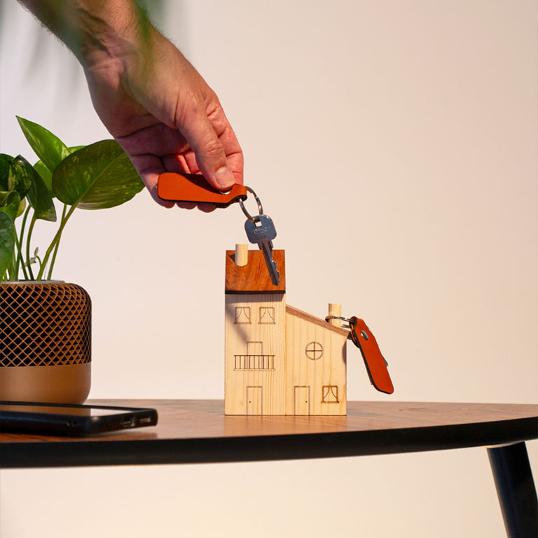 Fotografía de estudio del conjunto de peana o porta-llaveros con diseño de casitas de madera y su juego de llaveros de cuero grabados y cortados a láser